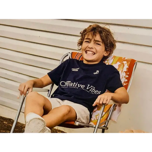 MAGLIETTA BLU RICAMATO PER BAMBINO di Retour, ragazzo sorridente seduto su sedia pieghevole