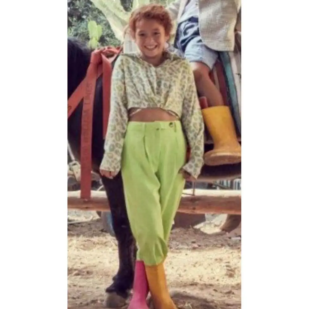 Bambina sorridente con camicia corta maculata bambina Y Clu abbinata a pantaloni verdi vita alta e stivali colorati