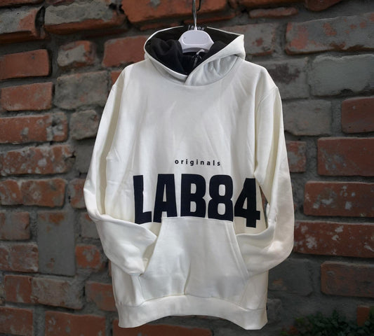 Felpa Bianca con cappuccio e tasca frontale per i ragazzi - Lab84 appesa a un muro di mattoni